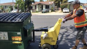 A TrashScouts employee in a safety vest using a Waste Caddy Lite to efficiently move a green dumpster in Oakland, CA.