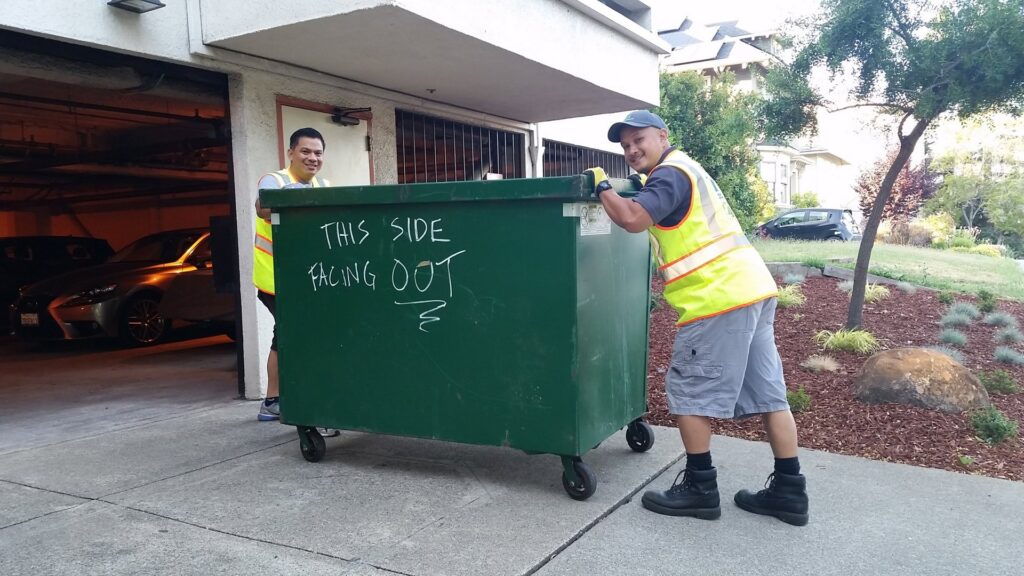 Two TrashScouts employees in safety vests working together to move a large green dumpster in Oakland, CA.