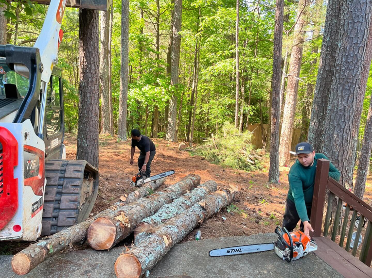 Two tree service workers using chainsaws to cut logs, with a skid steer nearby, for Ricardo's Tree Service in Woodstock, GA.