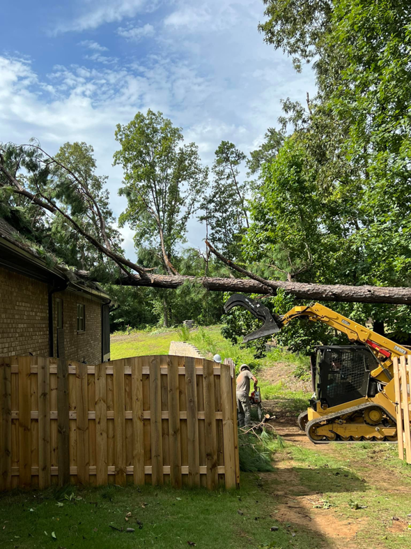 Workers use a skid steer loader with a grapple to remove a fallen tree near a house and fence for Ellison's Tree Service in Northport, AL.