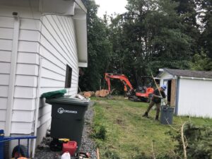 A tree service worker raking debris with an excavator and large logs in the background, showing cleanup by The Honest Arborist in Everett, WA.