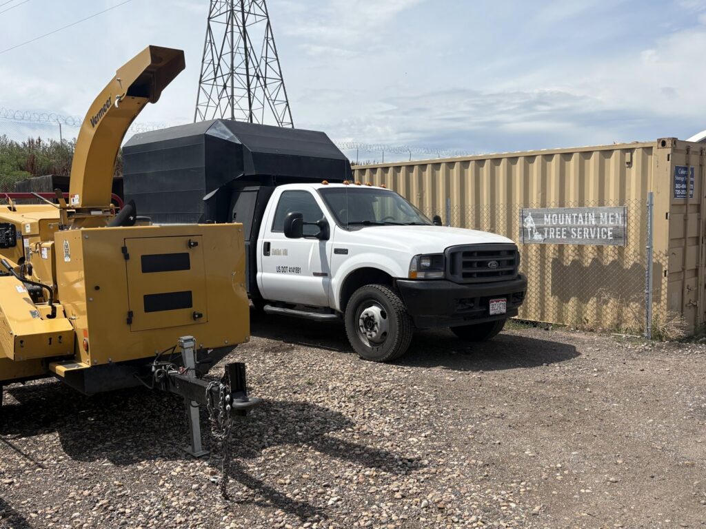 A Mountain Men Tree Service truck and wood chipper parked next to a fence with the business sign in Denver, CO.