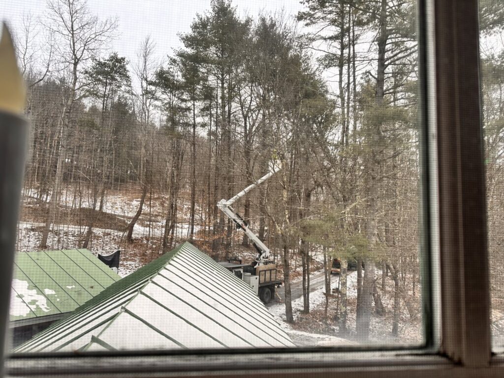 A tree service truck with a boom lift working on trees in a snowy, wooded area, seen from a window, by Teacher's Tree Service in South Burlington, VT.