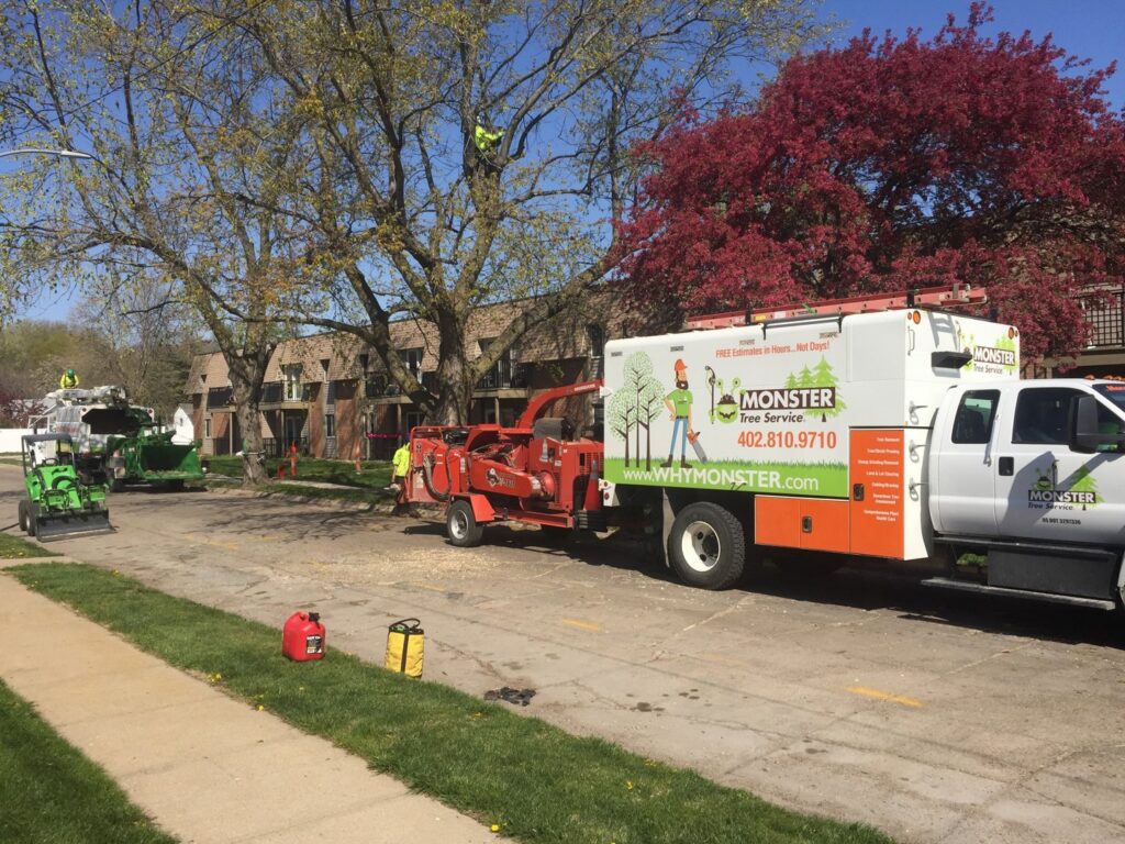 A Monster Tree Service truck and wood chipper are on a residential street with an arborist working in a tree in Omaha, NE.