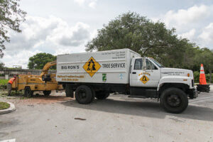 A Big Ron's Tree Service truck with a wood chipper and worker on a job site in Miami, FL.