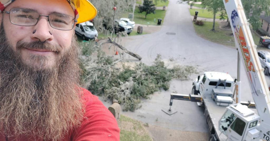 A tree service worker in a hard hat taking a selfie with a crane and tree debris in the background at a job site for Herrick's Tree Service in Jacksonville, FL.