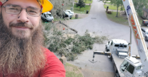 A tree service worker in a hard hat taking a selfie with a crane and tree debris in the background at a job site for Herrick's Tree Service in Jacksonville, FL.