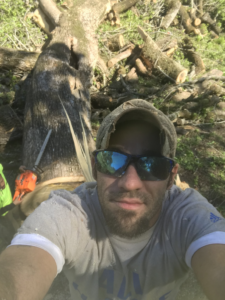 A tree service worker taking a selfie with a chainsaw and cut logs in the background, working for J.W. Tree Service in Montgomery, AL
