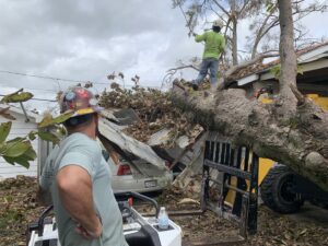 Tree service workers performing storm cleanup and tree removal with a skid steer near a damaged house for Tri-County Tree And Restoration in Jackson, MS.