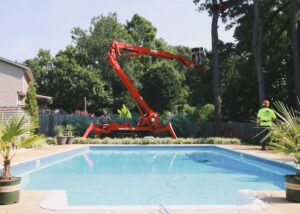 A worker in a spider lift trimming a tall pine tree over a swimming pool for Signature Tree Service in Greenville, SC.