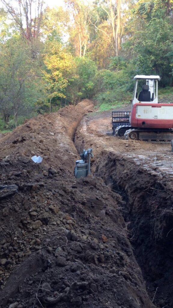 A long trench being dug by an excavator in a wooded area, showcasing excavation and site work by Horhut Inc. in Pittsburgh, PA.
