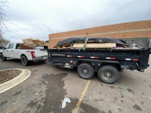 A white pickup truck pulling a dump trailer loaded with construction debris and covered by a tarp, handled by Hudson's Trash Removal, LLC in Great Falls, MT.