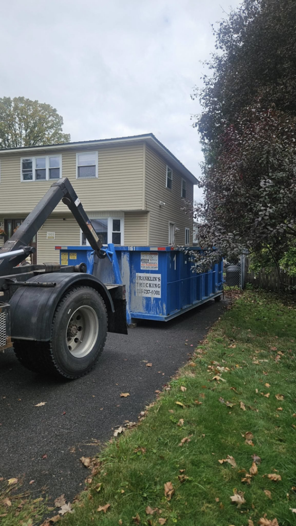 A Franklin's Disposal & Roll-Off Service truck delivering a blue dumpster to a residential driveway in Utica, NY.