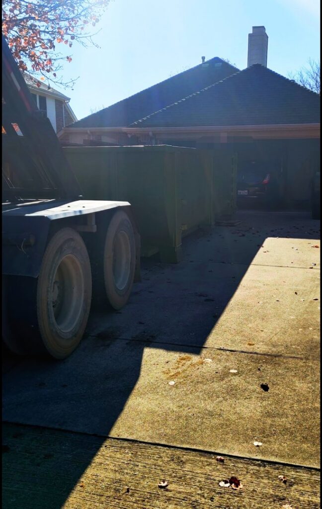 A FJD Waste Disposal and Recycling LLC truck delivering a green dumpster to a residential driveway for junk removal in Dallas, TX.