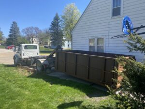 A NID Dumpster Rental truck delivering a large brown dumpster to a residential property in Nashua, IA.
