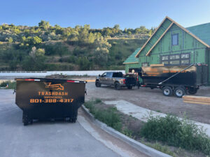 A TrashDash truck delivering two dumpsters to a construction site with a partially built house in West Haven, UT.