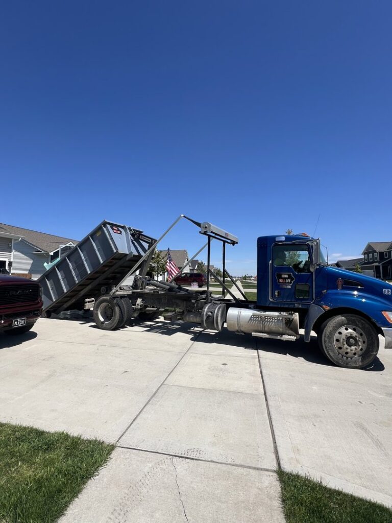 A blue roll-off truck actively delivering a grey dumpster onto a residential driveway for Iowa Roll Off in Ankeny, IA.