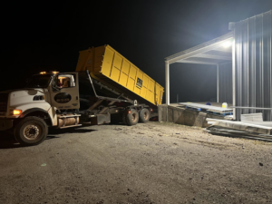 A large truck delivering a yellow roll-off dumpster at night for Tecumseh Rolloff Services in Moore, OK.