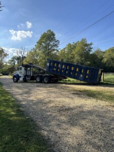 An AJ Carnevale Disposal truck deploying a blue dumpster on a gravel driveway in Hamilton, NJ