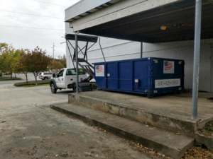 A truck deploys a blue roll-off dumpster at a commercial site for American Container Services, Inc in Columbia, SC.