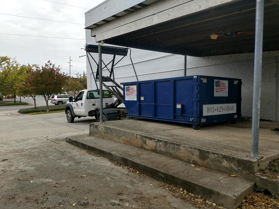 A truck deploys a blue roll-off dumpster at a commercial site for American Container Services, Inc in Columbia, SC.