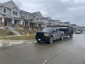 A black pickup truck hauling a dumpster trailer through a residential street for Waste Warriors Dumpster Rental of Des Moines, IA.