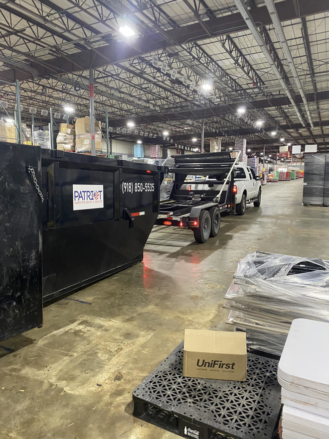 A truck hauling a large black dumpster on a trailer inside a warehouse for Patriot Dumpster Rental and Hauling in Tulsa, OK.