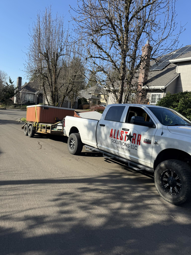 An AllStarr Solutions Hauling and Junk Removal truck hauling a large hot tub on a trailer down a residential street in Newberg, OR.