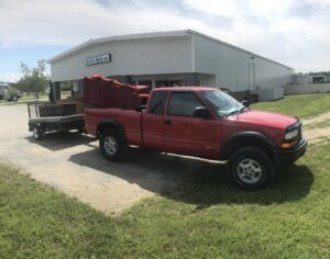 A red pickup truck hauling a sofa and other items on a trailer to Habitat ReStore by Hometown Hauling 802 in Milton, VT.