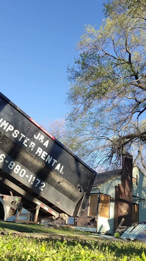 A J & A Haul Away & Dumpster Rentals truck lifting a black dumpster near a house in Derby, KS.