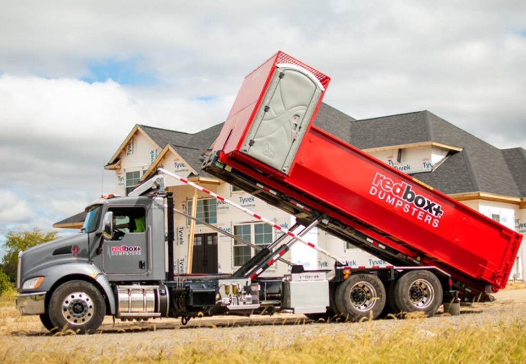Redbox+ truck lifting dumpster with portable toilet at construction site for redbox+ Dumpsters of Greater Austin, Austin, TX