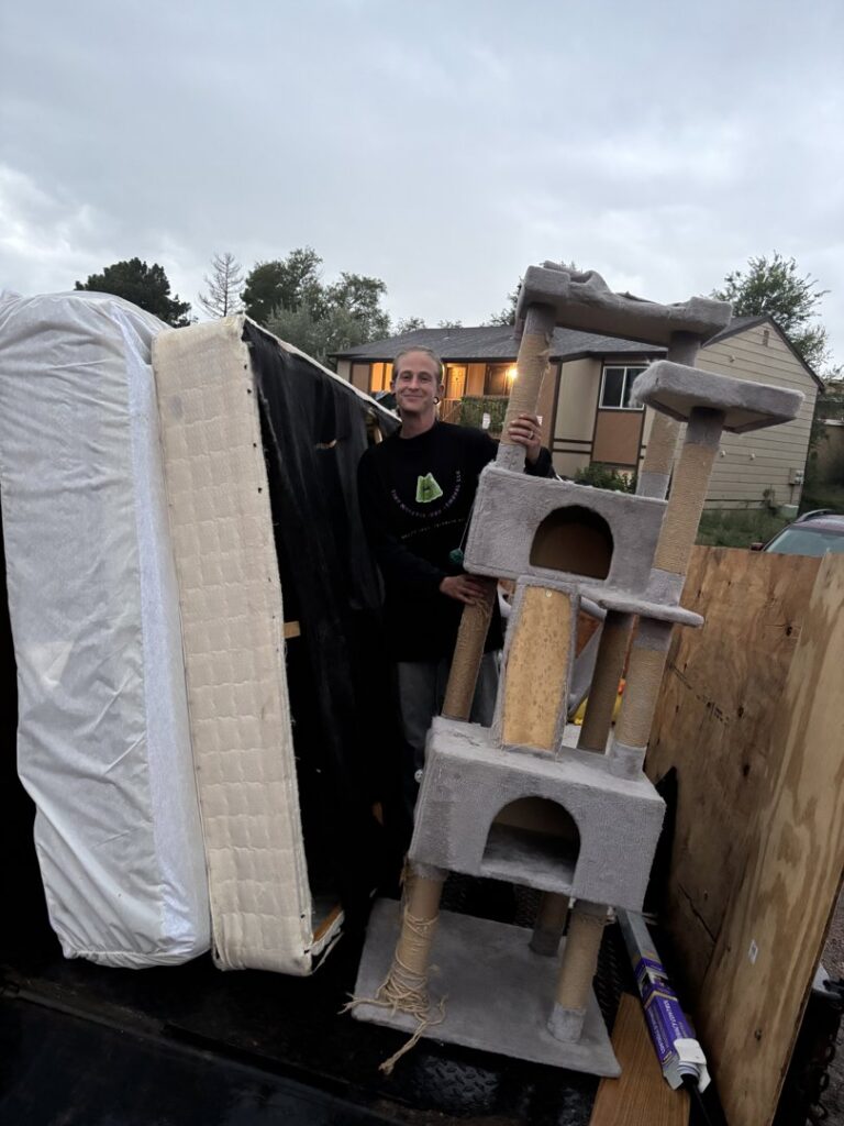 A worker from Tiny Monster Junk Removal LLC in Denver, CO, standing next to a truck loaded with mattresses and a cat tree.