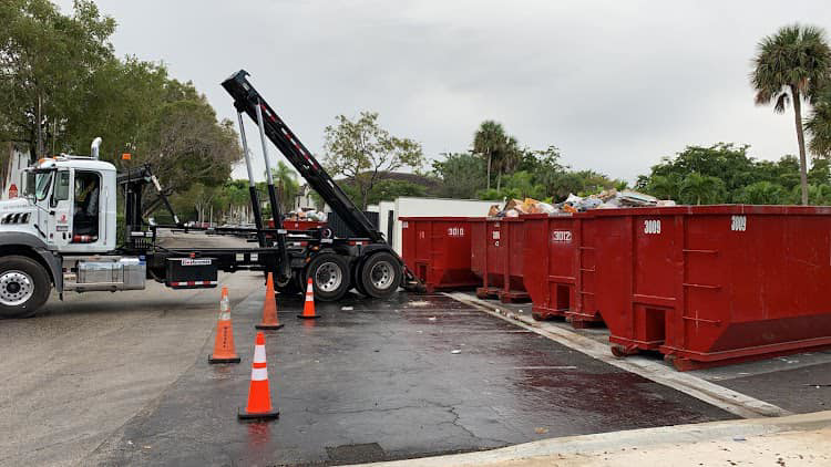 A hook-lift truck with multiple red dumpsters, some filled with junk, for Dumpster Rental Florida in West Park, FL.