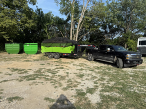 A truck pulling a covered dumpster trailer with two empty dumpsters in the background for Proper Dumpster Rentals in Kansas City, KS.