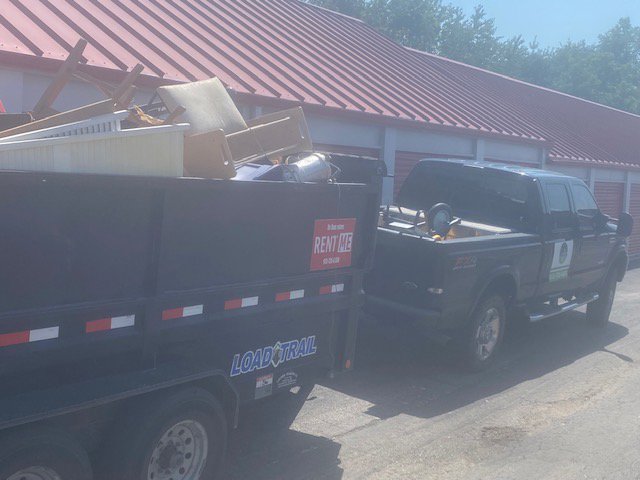 A pickup truck pulling a dump trailer filled with junk, parked outside storage units by D's Dumpster Rentals, Overland Park, KS.