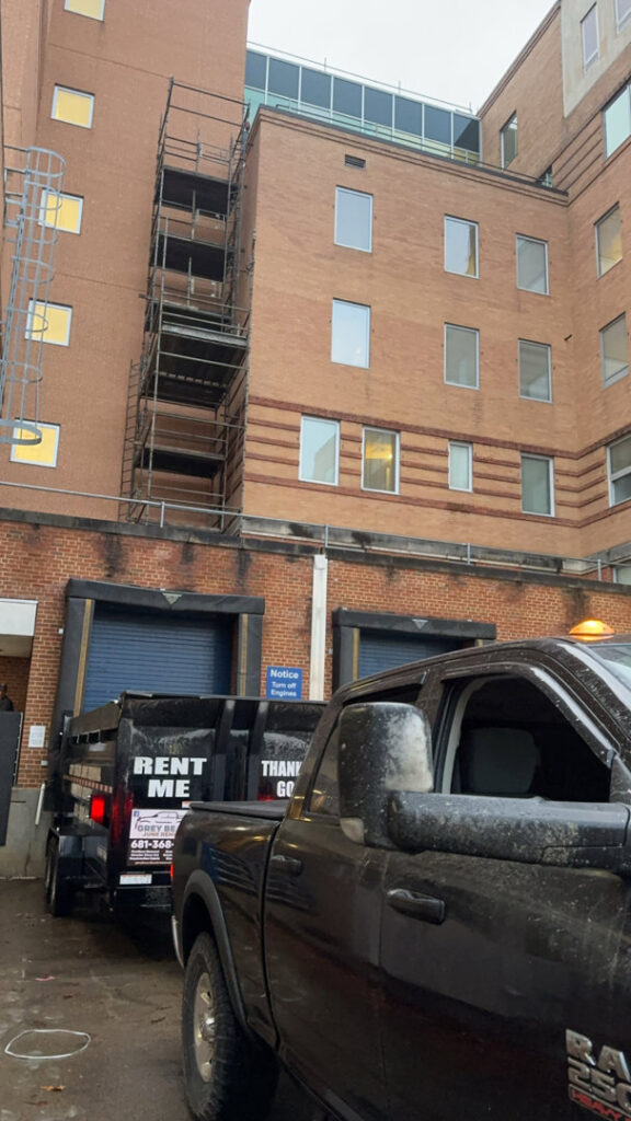 A Greybeard Dumpster Rentals truck towing a branded dumpster trailer in front of a building in Morgantown, WV.