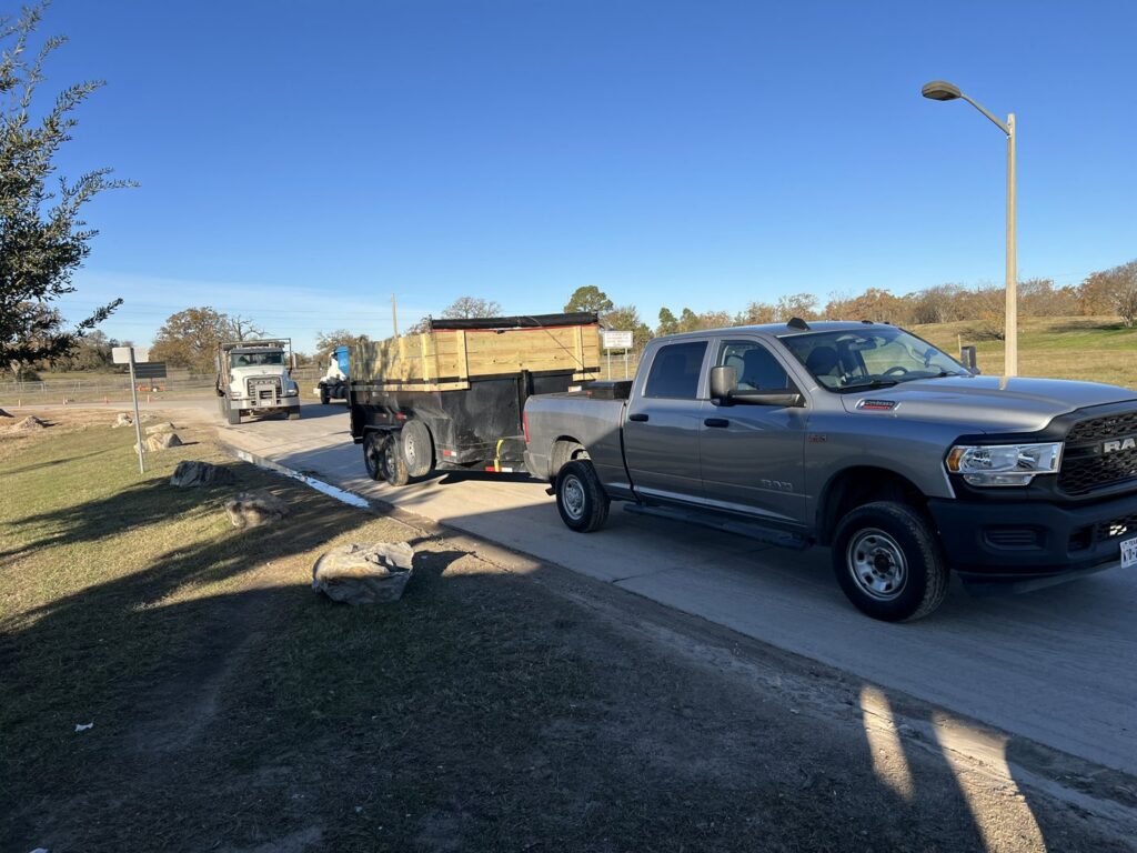 A pickup truck towing a dump trailer loaded with junk and debris down a road for Tidy Loaders Junk Removal in Houston, TX.