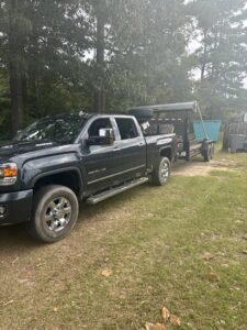 A pickup truck with a trailer and a blue dumpster in the background, ready for junk removal by Site Boss Dumpster Rentals in Raeford, NC.
