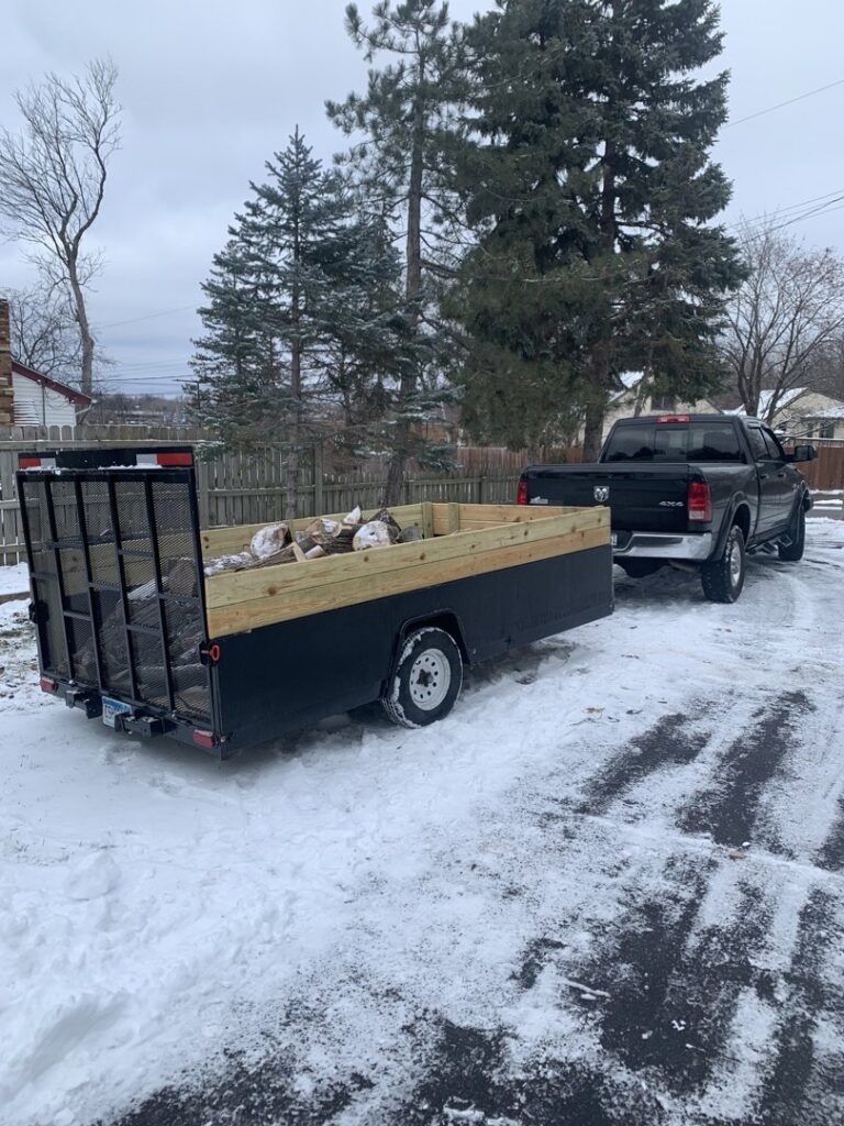 A truck and trailer loaded with logs and tree debris in a snowy driveway, handled by Mr. Groundhog Disposal and Services in Bloomington, MN.