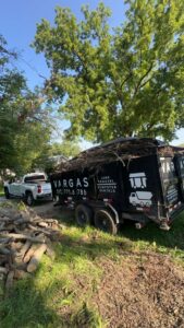 A Vargas Junk Removal & Dumpster Rentals truck and trailer next to a large pile of logs and branches for yard waste removal in Urbandale, IA.