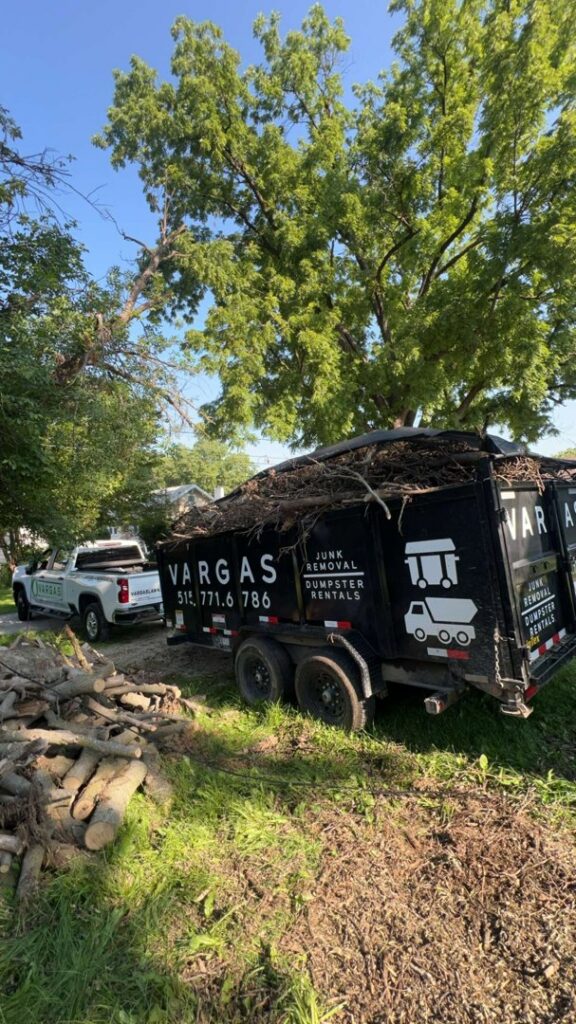 A Vargas Junk Removal & Dumpster Rentals truck and trailer next to a large pile of logs and branches for yard waste removal in Urbandale, IA.