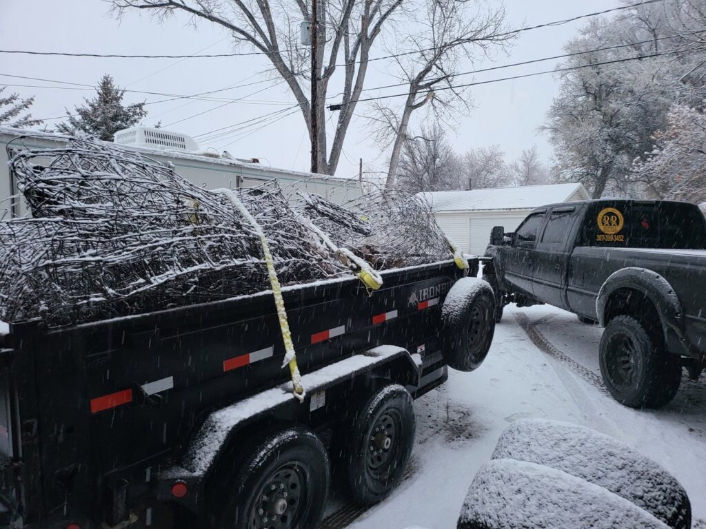 A pickup truck pulling a dump trailer filled with tangled yard waste and branches in the snow by Rubble Removers LLC in Riverton, WY.