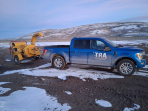 A blue TRA, Teton Rope Access and Services LLC pickup truck towing a wood chipper in a snowy landscape in Alpine, WY.