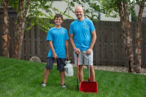 A Turd Terminators team member and assistant holding a pooper scooper and sprayer in a backyard in Omaha, NE.