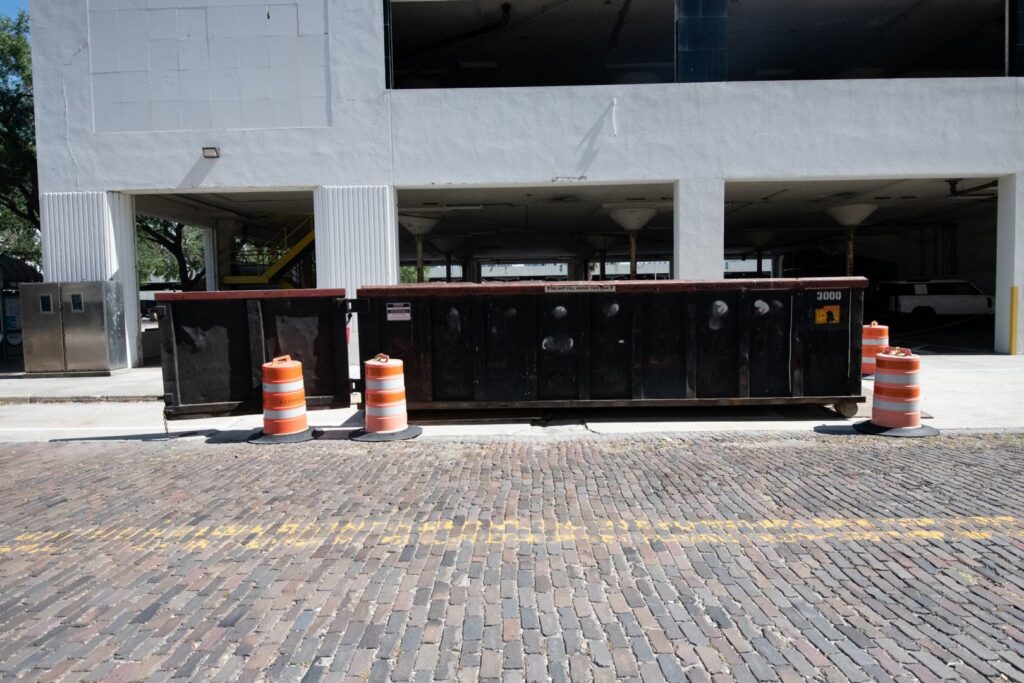 Two black roll-off dumpsters with orange traffic cones on a brick road, available from Waste Master Dumpster Rental Orlando, FL.