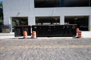 Two black roll-off dumpsters with orange traffic cones on a brick road, available from Waste Master Dumpster Rental Orlando, FL.