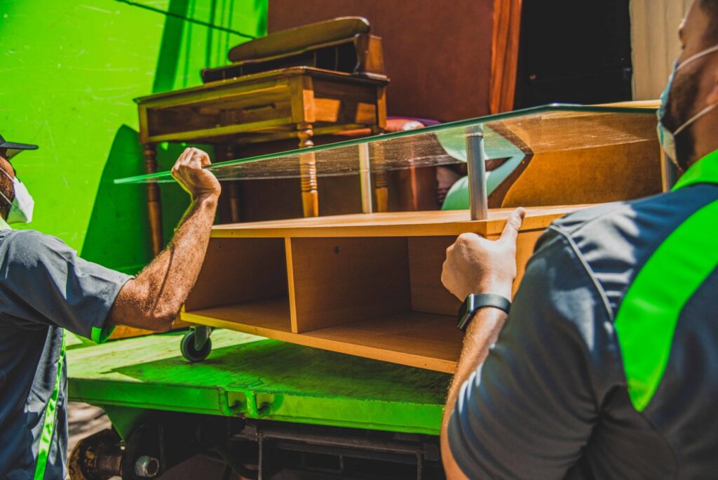 Two Junkluggers employees loading a wooden cabinet into a green junk removal truck for The Junkluggers in Hicksville, NY.