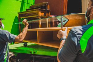 Two Junkluggers employees loading a wooden cabinet into a green junk removal truck for The Junkluggers in Hicksville, NY.