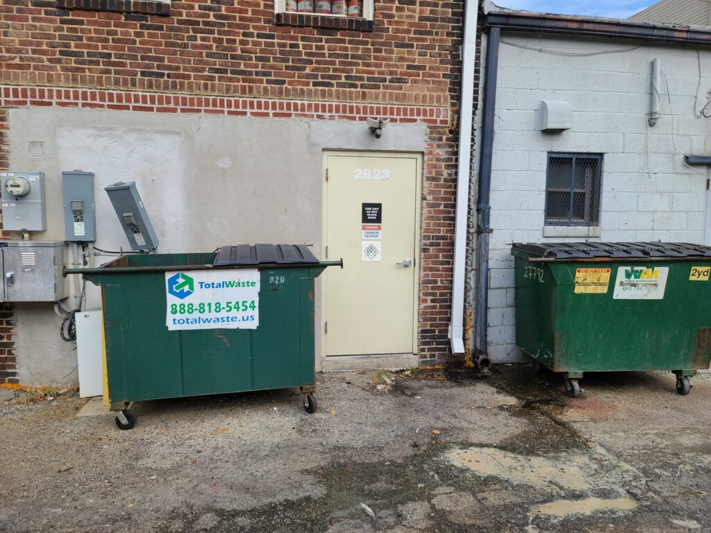 Two green dumpsters from Total Waste, one with an open lid, located behind a building in Baltimore, MD.