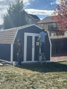Two handymen painting a shed in a backyard for Froese's Creative Concepts & Luxury Designs LLC in Denver, CO.